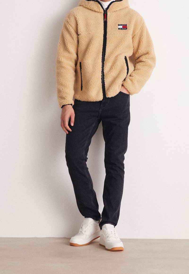 Persona che indossa una giacca in pile beige con zip e logo Tommy Jeans, jeans neri e sneakers bianche, in piedi con una mano nella tasca.