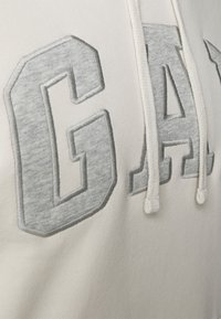 Gros plan sur un sweat à capuche beige avec de grandes lettres texturées grises "GA" et des cordons pendants à l'avant.