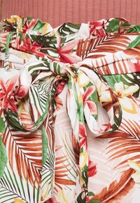 Ceinture enroulée florale présentant un mélange de motifs rouges, verts et jaunes sur un fond clair, avec un bord froncé et volants et une texture de tissu douce.