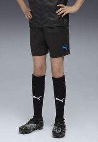 Črne športne kratke hlače z modrim logotipom Puma, v kombinaciji z zaščitnimi črnimi nogavicami do kolen, ki imajo bel logotip Puma, ter črnimi kopačkami z zelenimi poudarki.