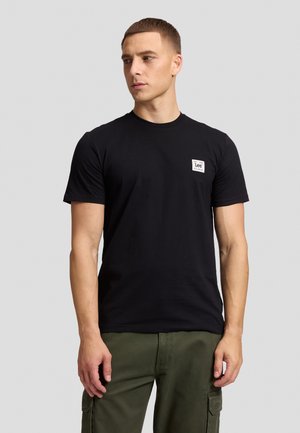 Homme portant un t-shirt noir à manches courtes avec un petit logo blanc sur la poitrine et un pantalon cargo vert olive, debout devant un fond gris.