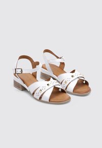 Sandalias de cuero blanco con tiras entrecruzadas, tachas doradas y un tacón bajo. Presentan una correa ajustable en el tobillo y una planta marrón.
