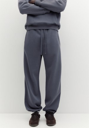 Homme portant un pantalon de survêtement ample gris foncé avec cordon de serrage et baskets marron foncé, debout les bras croisés.