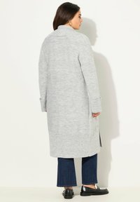 Dlouhý šedý cardigan s ribováním, s vysokým límcem, bočními rozparky a dlouhými rukávy, doplněný o tmavomodré džíny a černé boty.