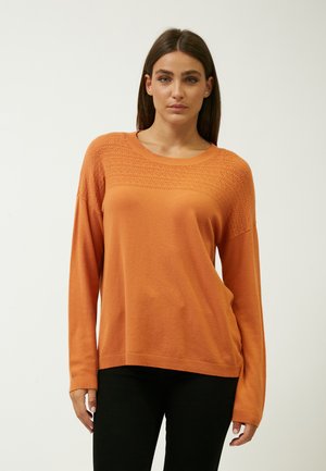 Oranger Pullover mit langen Ärmeln und einem strukturierten Strickmuster an der Oberseite, rundem Ausschnitt sowie gerippten Bündchen und Saum. Darunter werden schwarze Hosen getragen.