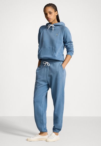 Mėlyna medvilnė hoodie su raišteliu ir priekinėmis kišenėmis, derinama su atitinkamomis jogging kelnėmis. Ypatybės: dirižiai ant rankovių ir laisvas siluetas. Baltos sportbačiai.