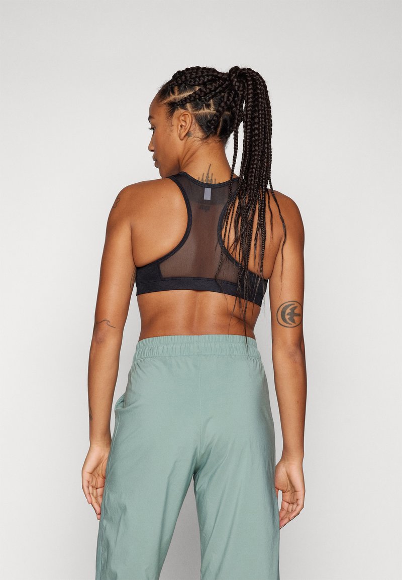 Reggiseno sportivo nero con schiena in rete, abbinato a pantaloni verdi chiari con cordino. Il reggiseno presenta un design elegante e una silhouette aderente.