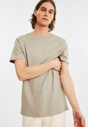 Lys beige kortærmet t-shirt i blødt stof med rund halsudskæring, afslappet pasform og ensartet design, båret af en person, der omfavner stoffet.