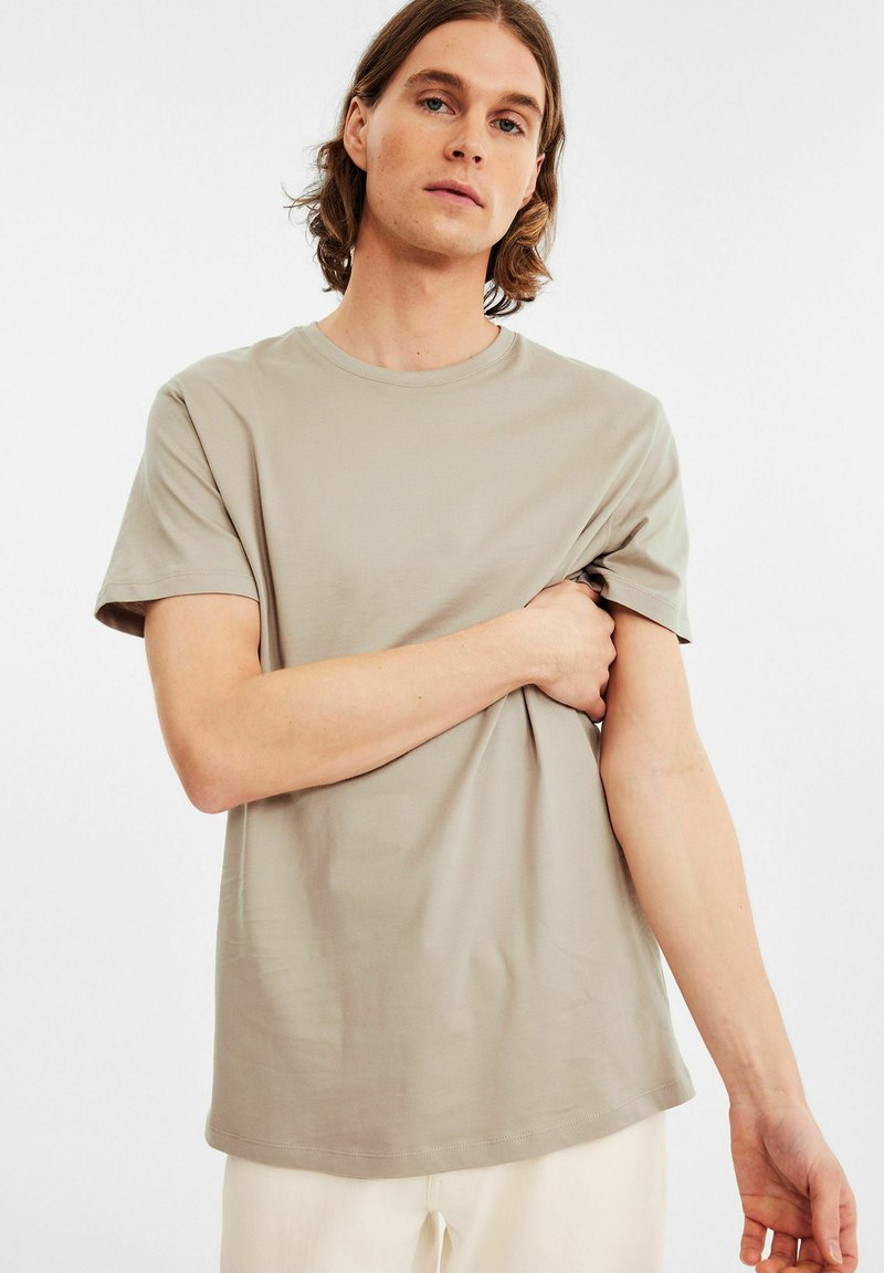 Lys beige kortærmet t-shirt i blødt stof med rund halsudskæring, afslappet pasform og ensartet design, båret af en person, der omfavner stoffet.