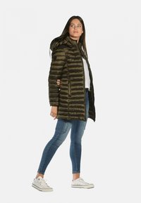 Manteau long rembourré vert foncé avec capuche, doté de rayures horizontales. Porté avec un jean bleu et des baskets blanches, un look décontracté.