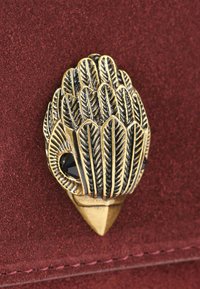 Ornement en métal doré avec un motif de feuille et une accent en pierre noire, attaché à un fond texturé bordeaux.