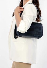 Czarna torebka o fakturze z teksturowanej skóry, z gładkim wykończeniem, z krótkim paskiem do noszenia na krzyż i ozdobnymi frędzlami, trzymana przy białym żakiecie.