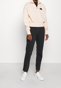 Jasno beżowa bluza z zamkiem błyskawicznym, z prążkowanymi mankietami i dołem, w zestawie z dopasowanymi czarnymi dżinsami i białymi trampkami. Logo CK na piersi.