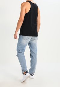 Mężczyzna w czarnej koszulce bez rękawów, jasnoniebieskich dżinsach i białych trampkach Adidas idzie w kierunku białego tła.