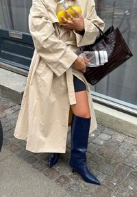 Trench coat beige, stivali al ginocchio blu con stampa a coccodrillo e punta affusolata, borsa nera strutturata, mentre tiene un limone e una bottiglia d'acqua.