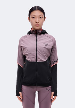 Une femme porte une veste de sport zippée mauve et noire, assortie à un legging, sur un fond uni et clair.
