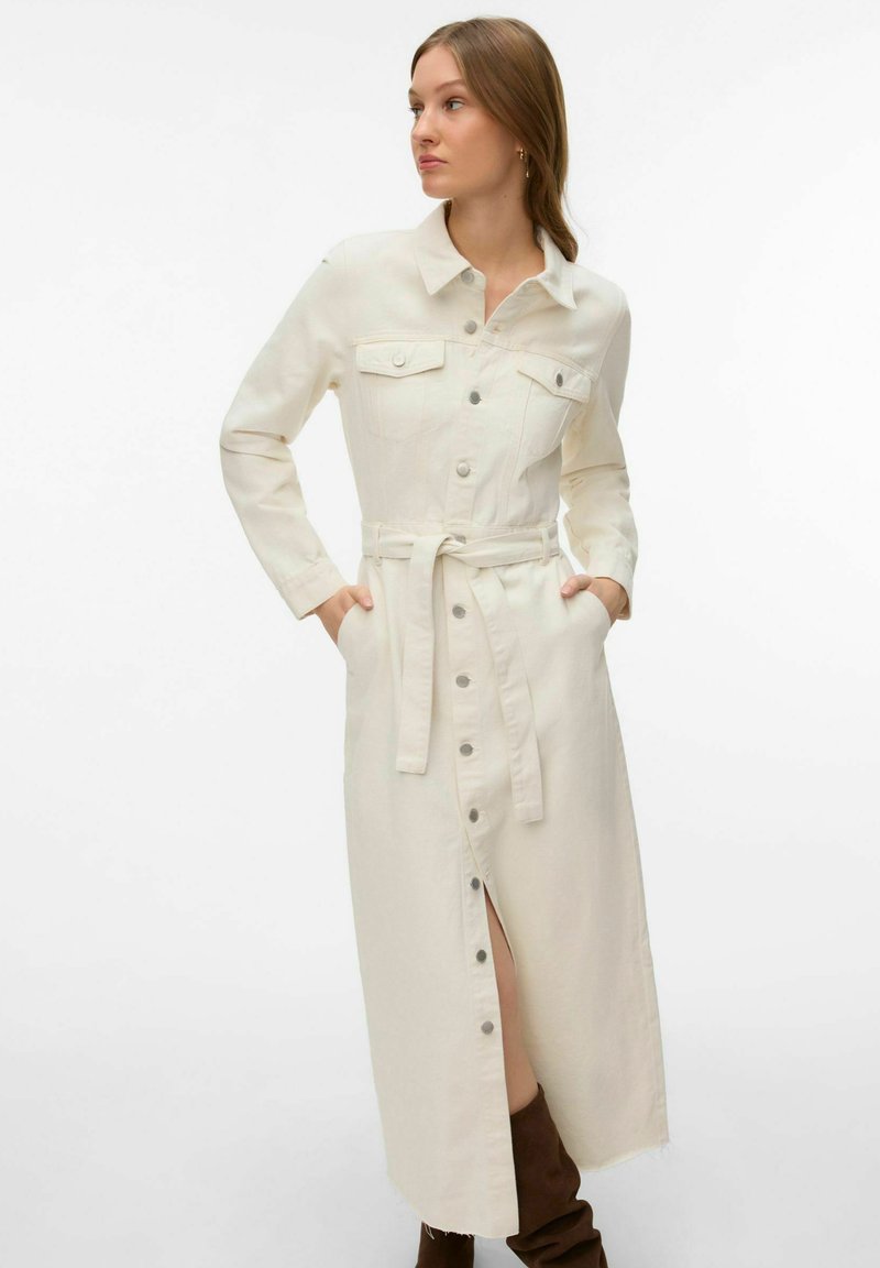 Femme en robe longue crème boutonnée avec poches et ceinture, mains dans les poches, regardant vers la droite, portant des bottes marron foncé.