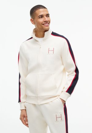 Junger Mann lächelt, trägt weiße Jacke mit Reißverschluss und Hosen mit roten und marineblauen Streifen sowie Logo-Patches auf Brust und Oberschenkel.