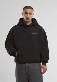 Schwarzer Kapuzensweatshirt mit einer Fronttasche und dem Text "VERBOTEN" auf der Brust. Das Material wirkt weich und dick mit einer lockeren Passform.