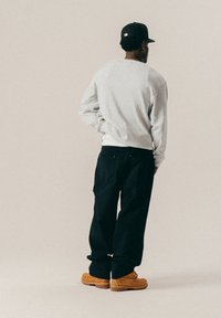 Mees musta mütsiga, halli dressipluusi, mustade pükste ja naturaalsest nahast saabastega, seisab seljaga pööratuna, üks käsi selja taga.