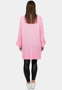 Overmaatse roze gebreide trui met wijde mouwen, gestructureerde stof, gedragen over zwarte leggings en witte sneakers, die een ontspannen silhouet laat zien.