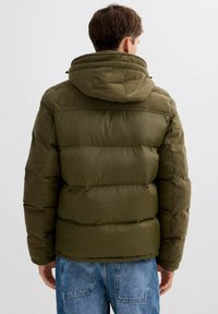 Chaqueta acolchada verde oliva con capucha, con un diseño acolchado que combina secciones lisas y detalles acanalados en los hombros y la capucha.