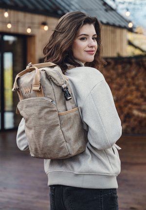 Beiger Stoff-Rucksack mit braunen Akzenten, verstellbaren Trägern und einer Vordertasche. Texturiertes Material mit einem lässigen Design, seitlich betrachtet.