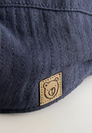 Tumesinist värvi kangas soonilise tekstuuriga, millel on nahast plaastriga karu logo. Plaaster on ruudukujuline õmmeldud äärtega.