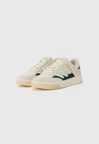 Beige sneakers with a textured, mesh upper and green accents. Features a thick rubber sole and a unique wavy design on the sides.