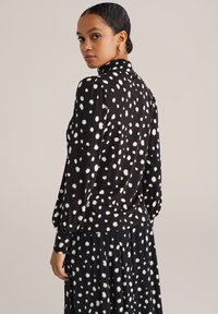 Kobieta ubrana w czarną bluzkę z długim rękawem i spódnicę w białe nieregularne kropki, patrząca przez ramię, z włosami związanymi do tyłu.