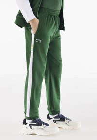 Zelene trenirke s bijelim bočnim trakama, elastičnom strukom i Lacoste logom. Uparene s bijelim i tamnoplavim tenisicama. Mekana, teksturirana tkanina.