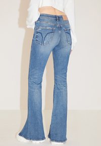 Jeans de mezclilla azul acampanados con un lavado medio, que presentan bolsillos traseros con costura delineada y un dobladillo sin rematar en la parte inferior.