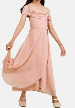 Fille portant une robe rose fluide à épaules dénudées avec des manches transparentes et des ornements en perles, tenant la jupe en se tenant debout.