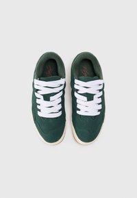 Zielone zamszowe sneakersy z białymi sznurówkami, charakteryzujące się teksturowanym wzorem i kontrastującą gumową podeszwą. Widoczny minimalistyczny styl i plecione akcenty.