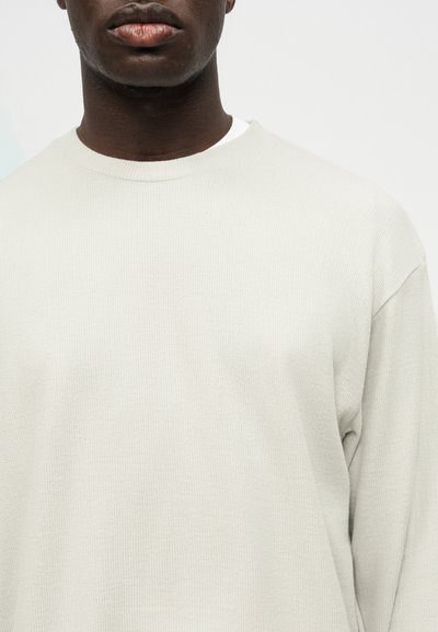 Κοντινό πλάνο ενός ατόμου που φοράει ανοιχτό μπεζ ριμπ πουλόβερ με μακριά μανίκια πάνω από ένα λευκό εσώρουχο, εμφανιζόμενο από το στήθος και πάνω.