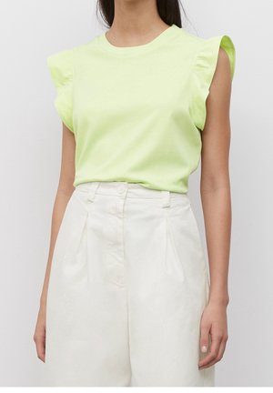 Femme portant un haut volanté sans manches vert clair rentré dans un pantalon blanc plissé taille haute, debout devant un fond uni.