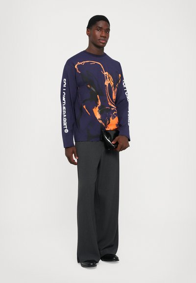 Navy sweatshirt with orange graphic design, long sleeves, and white text on the sleeves, paired with gray wide-leg trousers and a black clutch.