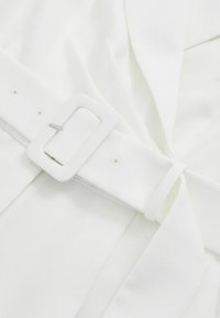 Balts audums ar gludu tekstūru, kam ir josta ar taisnstūrveida baltu sprādzi, aizdarināta ar sudraba detaļām, un salocītu drapējuma detaļu.