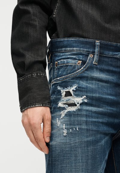 Main reposant à côté, portant une veste en denim foncé et un jean bleu avec un tissu usé et déchiré sur la cuisse.