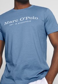 Camiseta de algodón azul con mangas cortas, que presenta el texto impreso en blanco "Marc O'Polo EST. EN ESTOCOLMO" en el pecho. Textura suave.