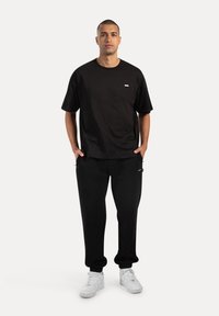 T-shirt nera a maniche corte con un piccolo logo bianco sul petto, abbinata a pantaloni jogger neri e scarpe da ginnastica bianche. Tessuto liscio, vestibilità rilassata.