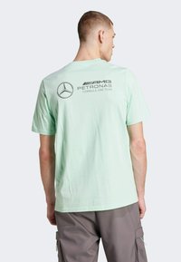 Camiseta verde menta con un gran logo de Mercedes y el texto "AMG PETRONAS FORMULA ONE TEAM" en la parte posterior. Mangas cortas, corte relajado.