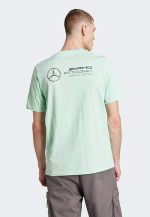 Majica mint zelene boje s velikim Mercedesovim logom i natpisom "AMG PETRONAS FORMULA ONE TEAM" na stražnoj strani. Kratki rukavi, opušteni kroj.