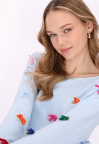 Maglione azzurro con applicazioni a fiocco 3D colorate in rosa, arancione, blu, verde e viola. Tessuto morbido e vestibilità rilassata.