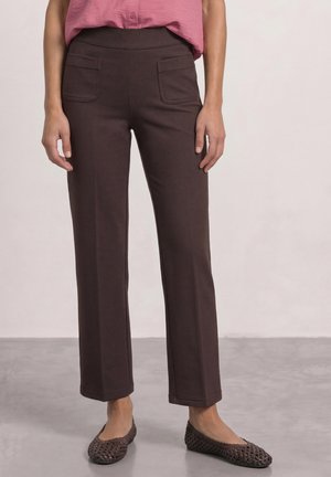 Frau trägt dunkelbraune gerade geschnittene Hose mit aufgesetzten Fronttaschen, rosa Hemd mit Knöpfen und braune geflochtene Ballerinas.