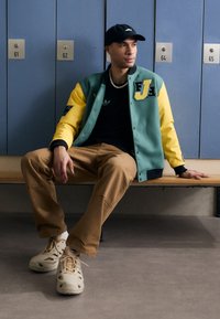Green and yellow varsity jacket with letter patches, black shirt, tan pants, and beige shoes. Posing on a bench in front of lockers.