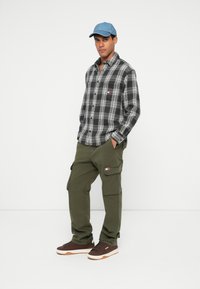 Juodas ir baltas languotas marškinėliai, alyvuogių spalvos kargo kelnės, mėlyna kepurė ir rudi sportbačiai. Išsiskiriantis logotipas ant marškinėlių ir kelnių kišenių.