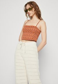 Narančasti crop top s uzorkom i tankim naramenicama, uparen s kremastim širokim hlačama teksture koje imaju horizontalne pruge.