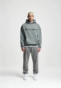 Felpa grigia con cappuccio con testo "2Y STUDIOS", abbinata a pantaloni della tuta grigi coordinati. Sneakers bianche chunky completano l'outfit.