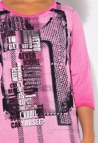 Top en tissu rose avec un design graphique noir comprenant du texte, des points et des motifs ; encolure ronde et manches trois-quarts.